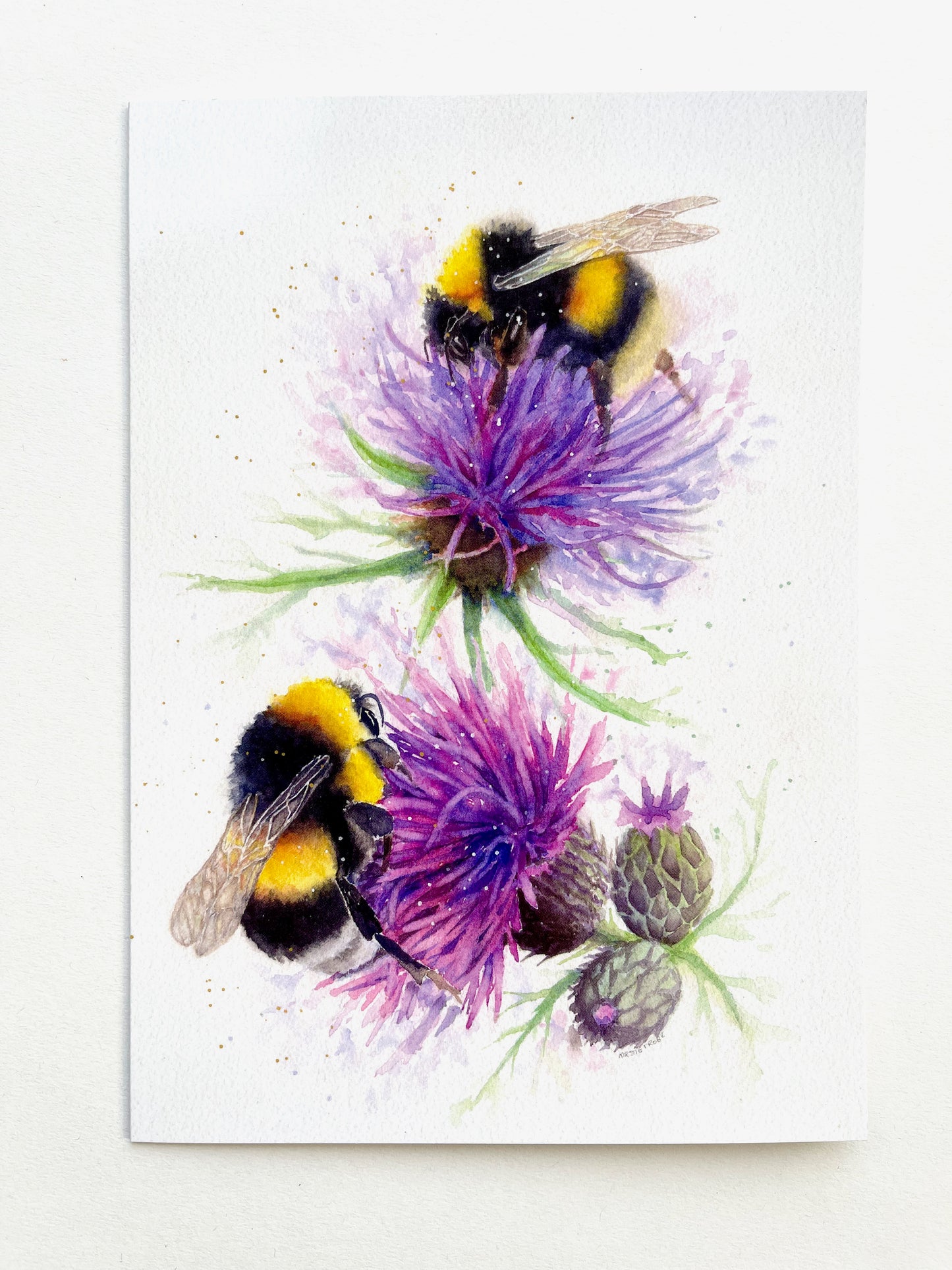 Two fluffy, fuzzy bumble bees feed on two thistles. The yellow and black of their fur contrasts with the purple pink of the thistle flowers. The bees are positioned one above the other in a portrait orientation